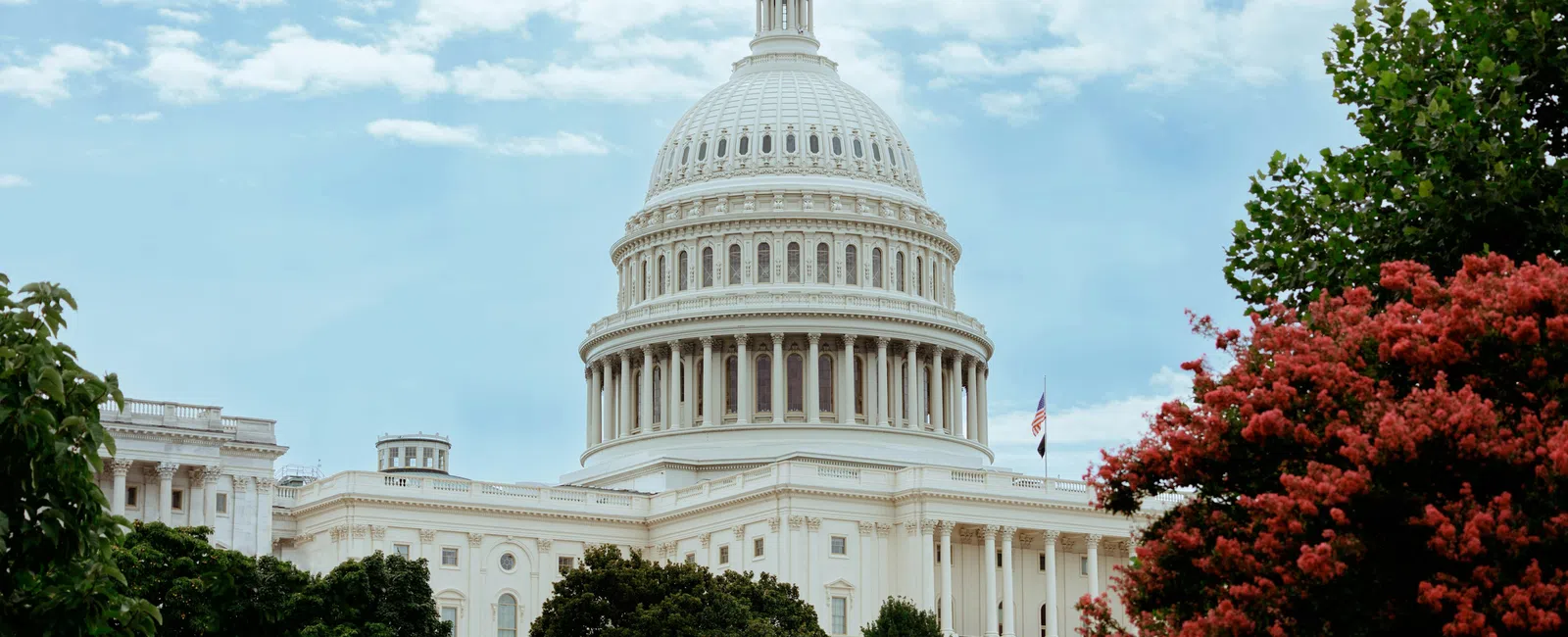 The United States Capitol building with its white dome surrounded by green and red foliage under a partly cloudy sky.