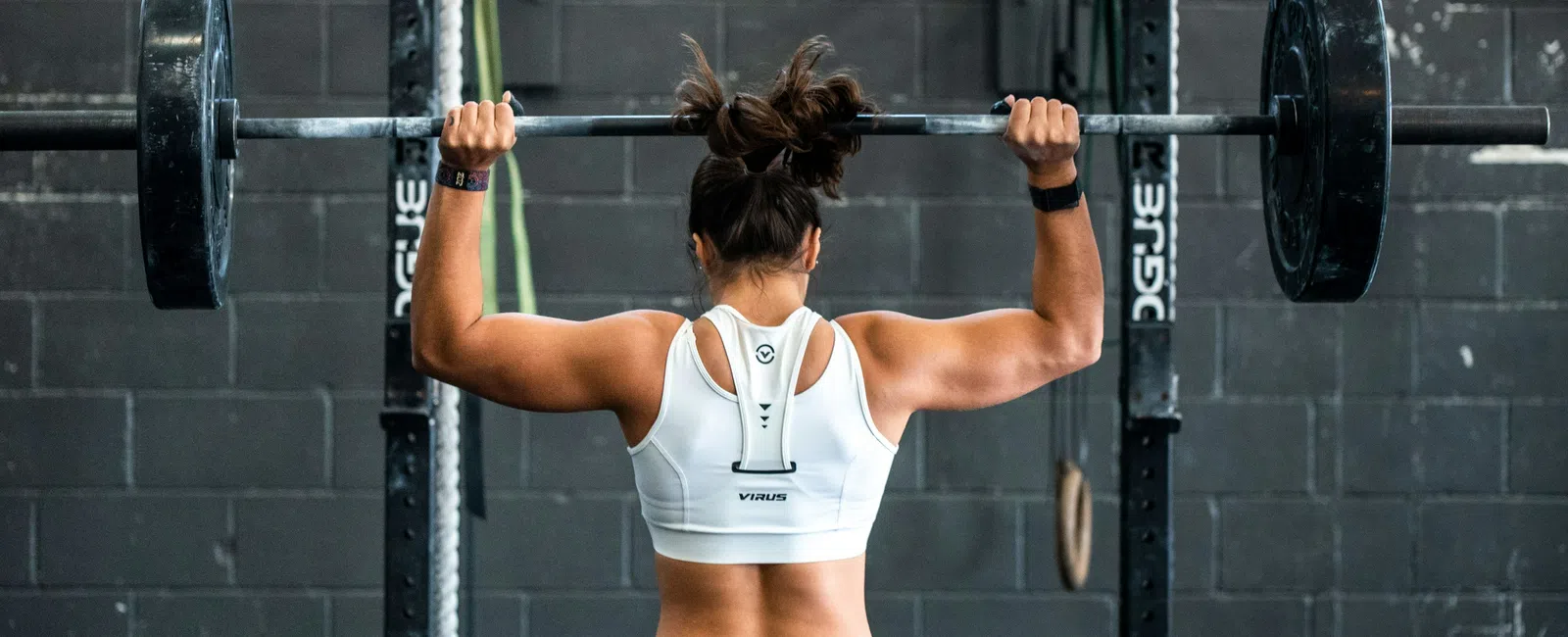 Woman lifting barbell weight in gym, seen from behind with muscular arms and white sports top