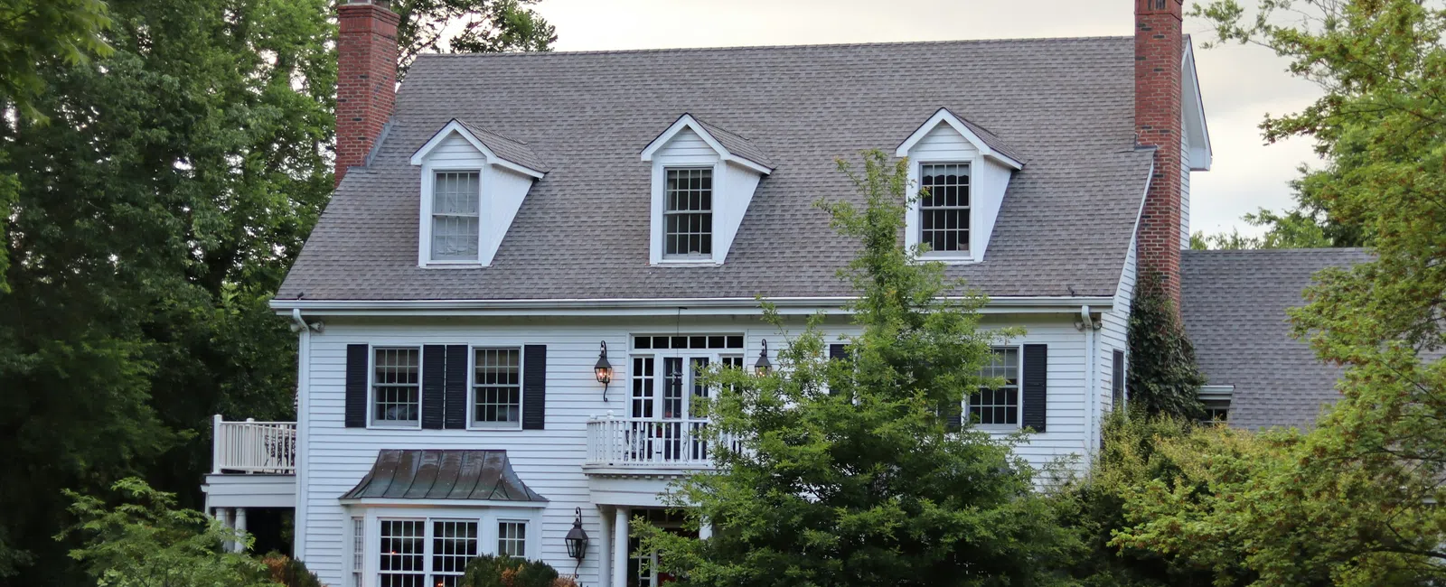 White two-story colonial house with black shutters, dormer windows, and lush green trees surrounding the lawn