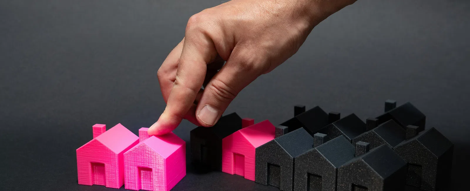 Hand placing a pink 3D printed house among black houses on a black surface with studio lighting.