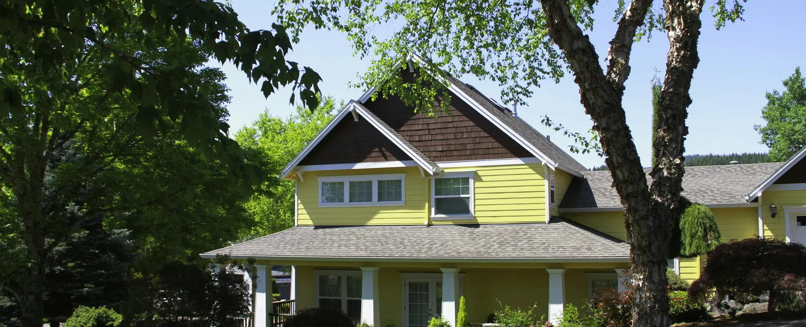 Sunny yellow two-story house with porch surrounded by lush green trees and a well-kept lawn on a bright day
