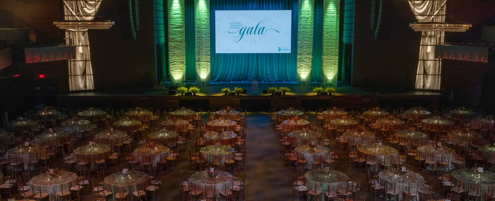 Elegant gala event setup with round tables, chairs, lit candles, and a large stage with green uplighting and a screen.