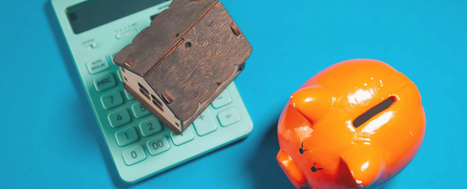 Orange piggy bank, wooden house model on calculator, blue background representing savings and budgeting.
