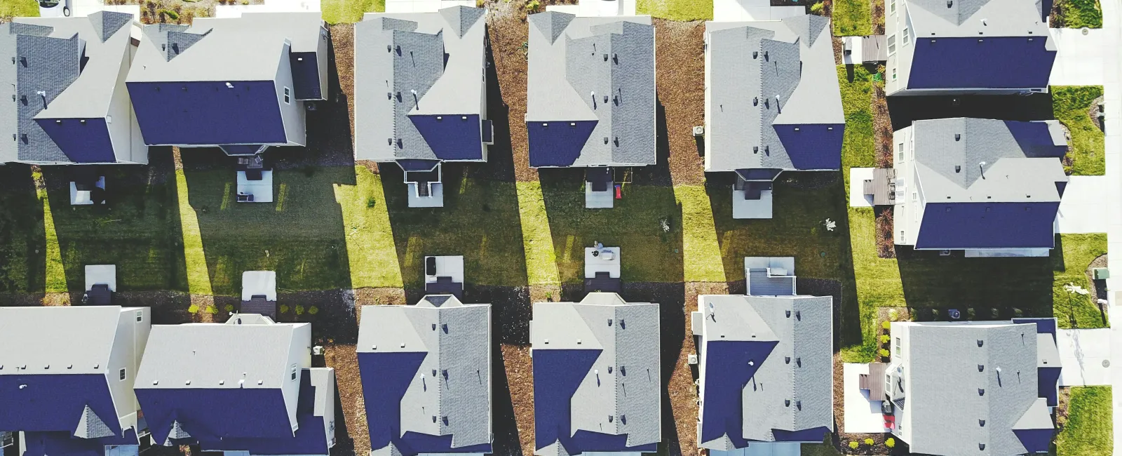 Aerial view of a suburban neighborhood showing rows of houses with driveways, lawns, and parked cars.