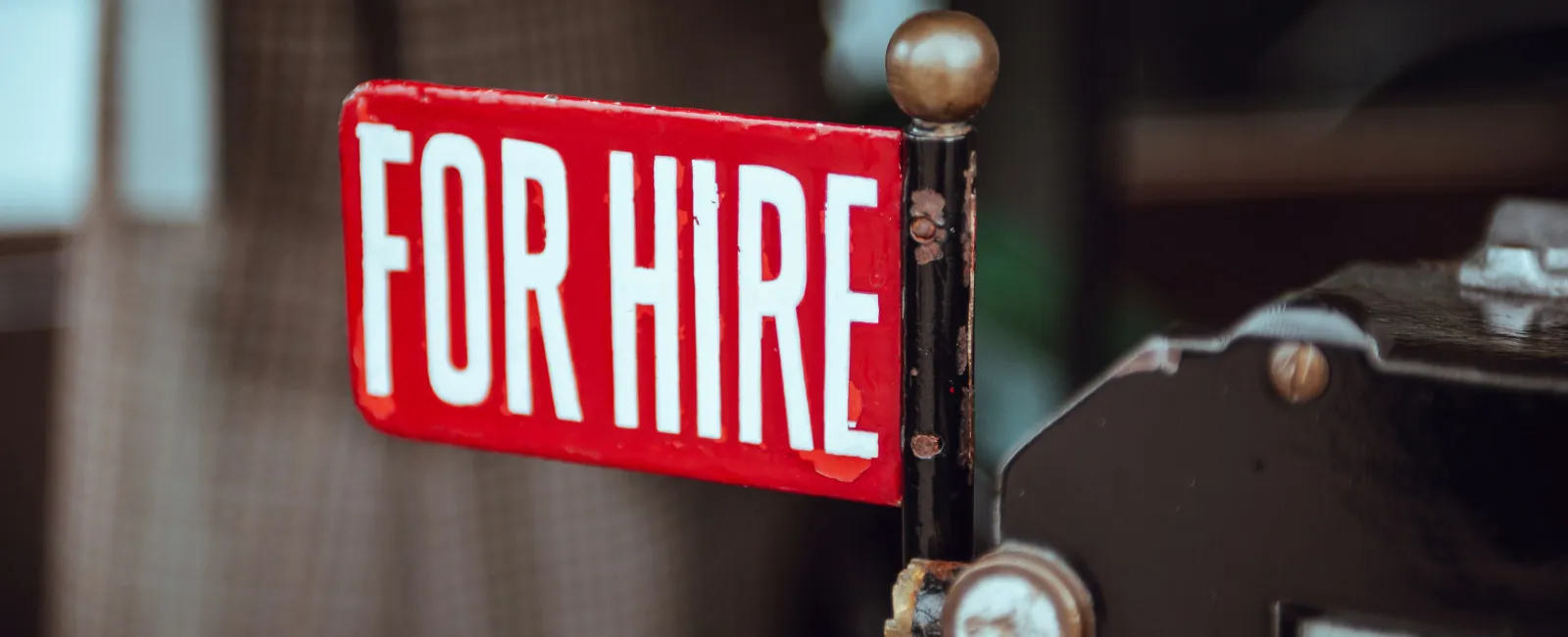 Red and white vintage 'For Hire' sign mounted on a classic vehicle against a blurred background.
