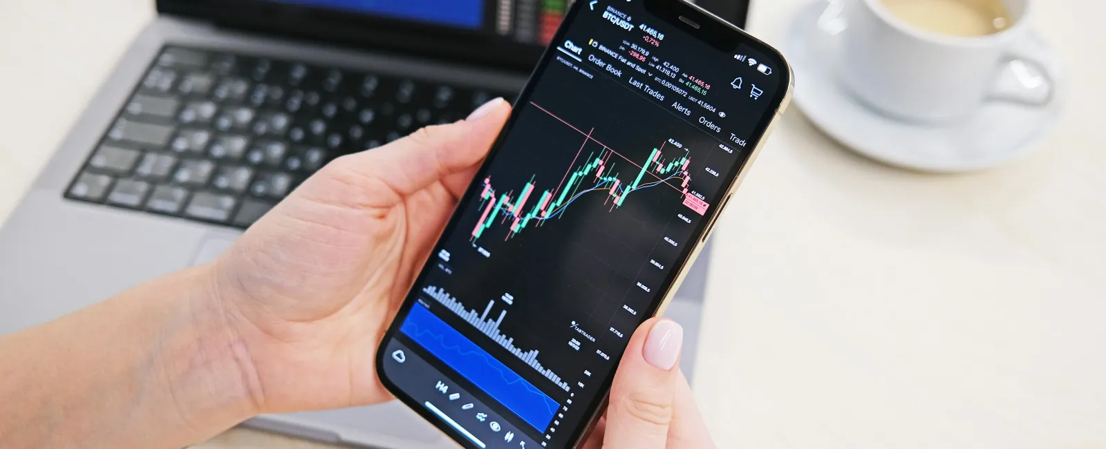 Person holding smartphone showing stock market charts with laptop and coffee cup in the background