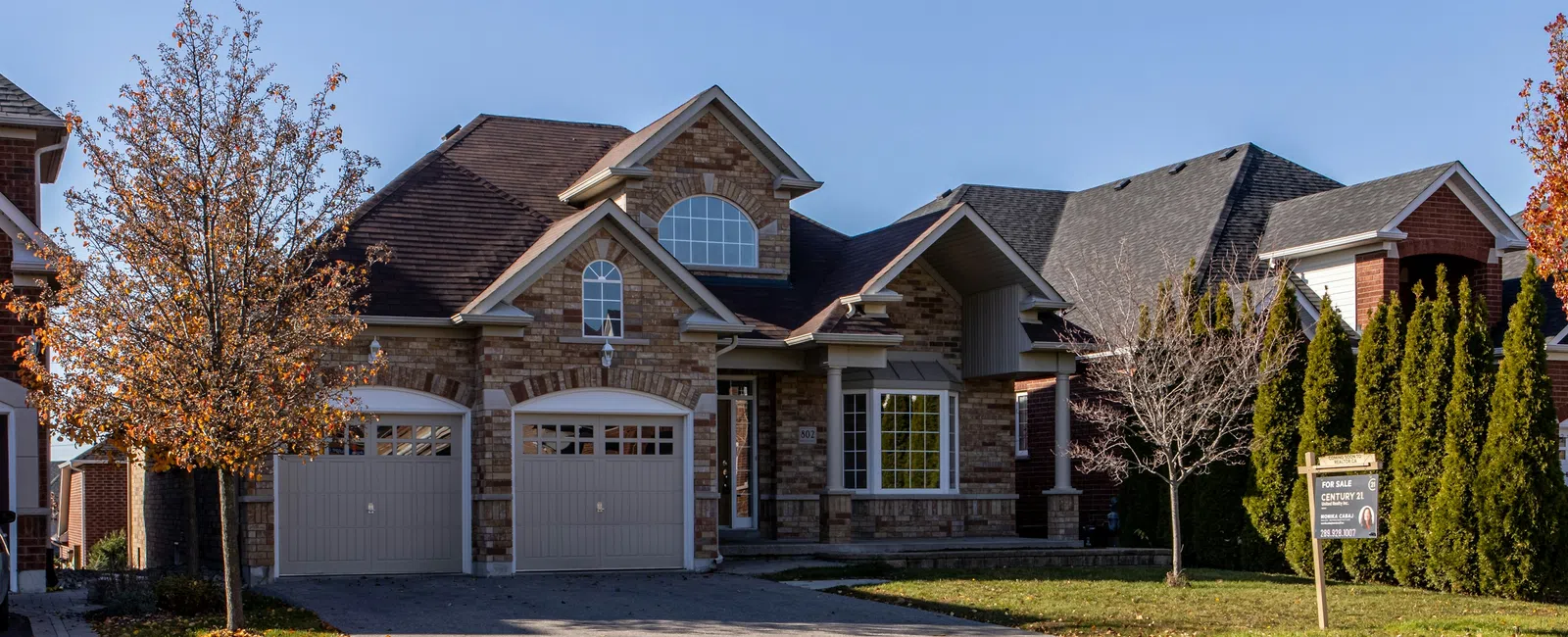 Two-story brick house with double garage doors and autumn trees under a clear blue sky.