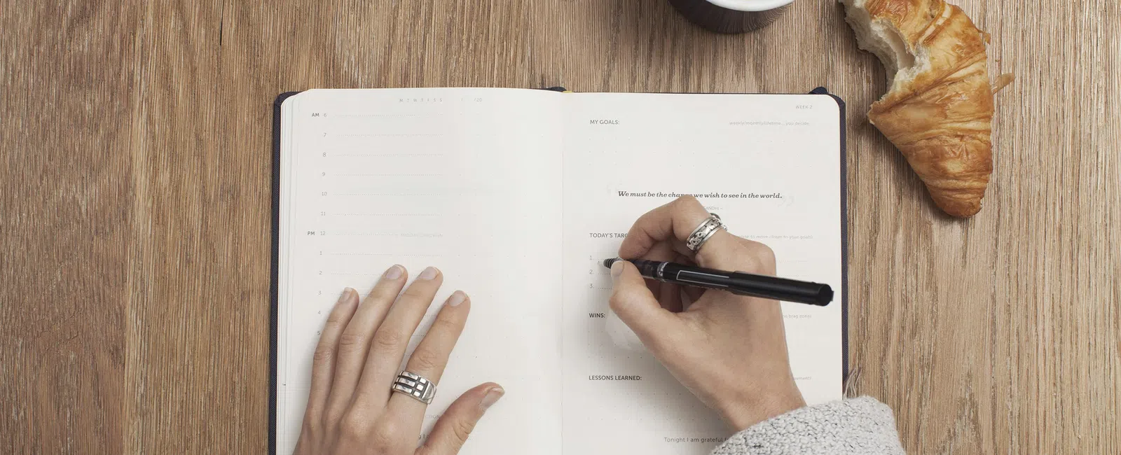 Person writing in a planner with a cup of coffee and a croissant on a wooden table.