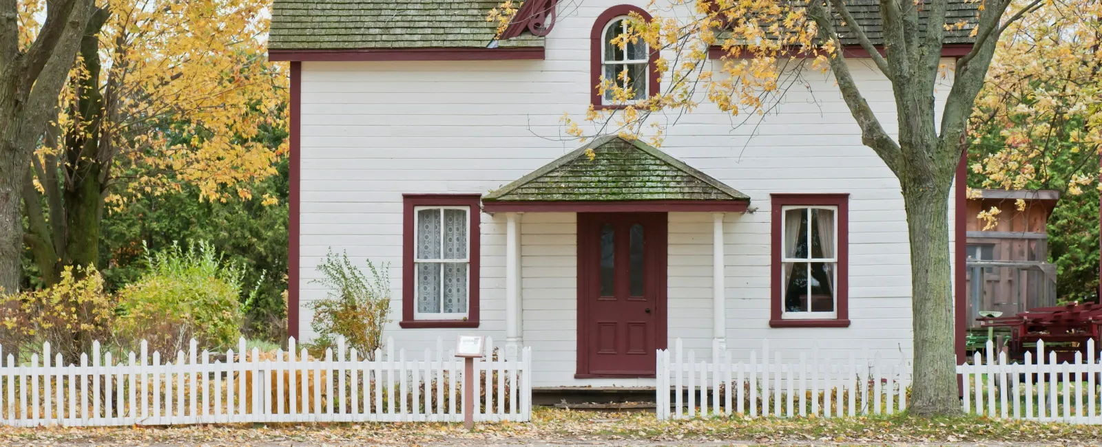 White house with red trim and a picket fence surrounded by autumn trees and fallen leaves.