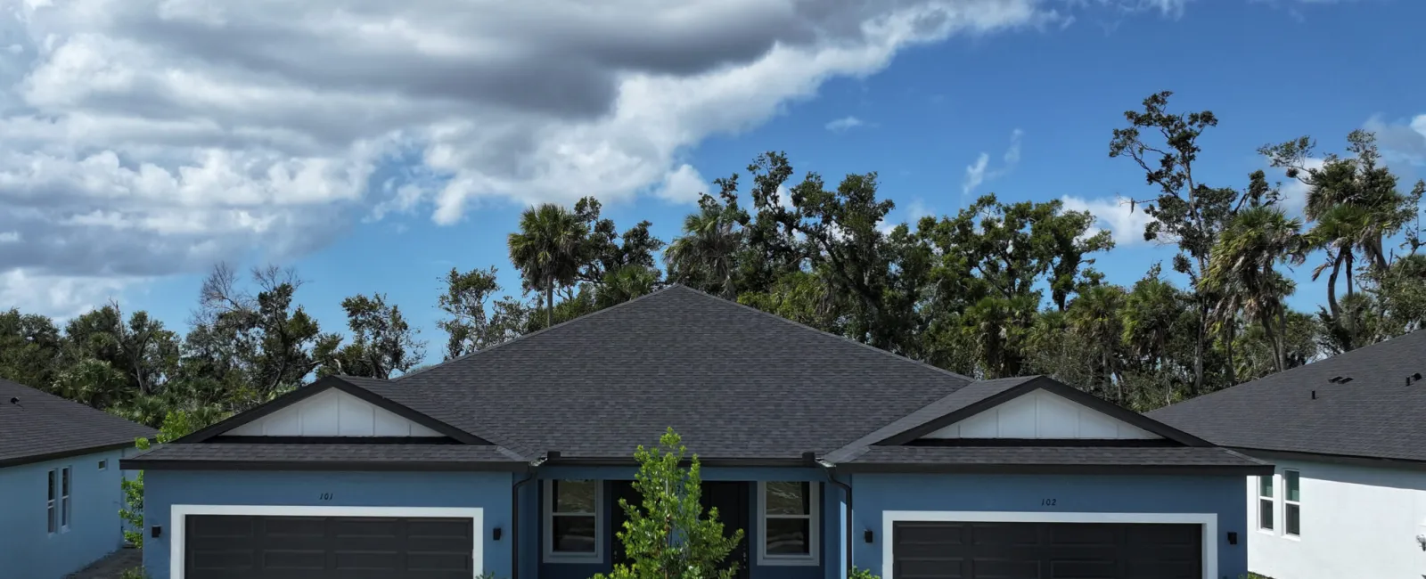 Modern blue duplex home with black roofs, large garages, parked black car, greenery, and cloudy blue sky.