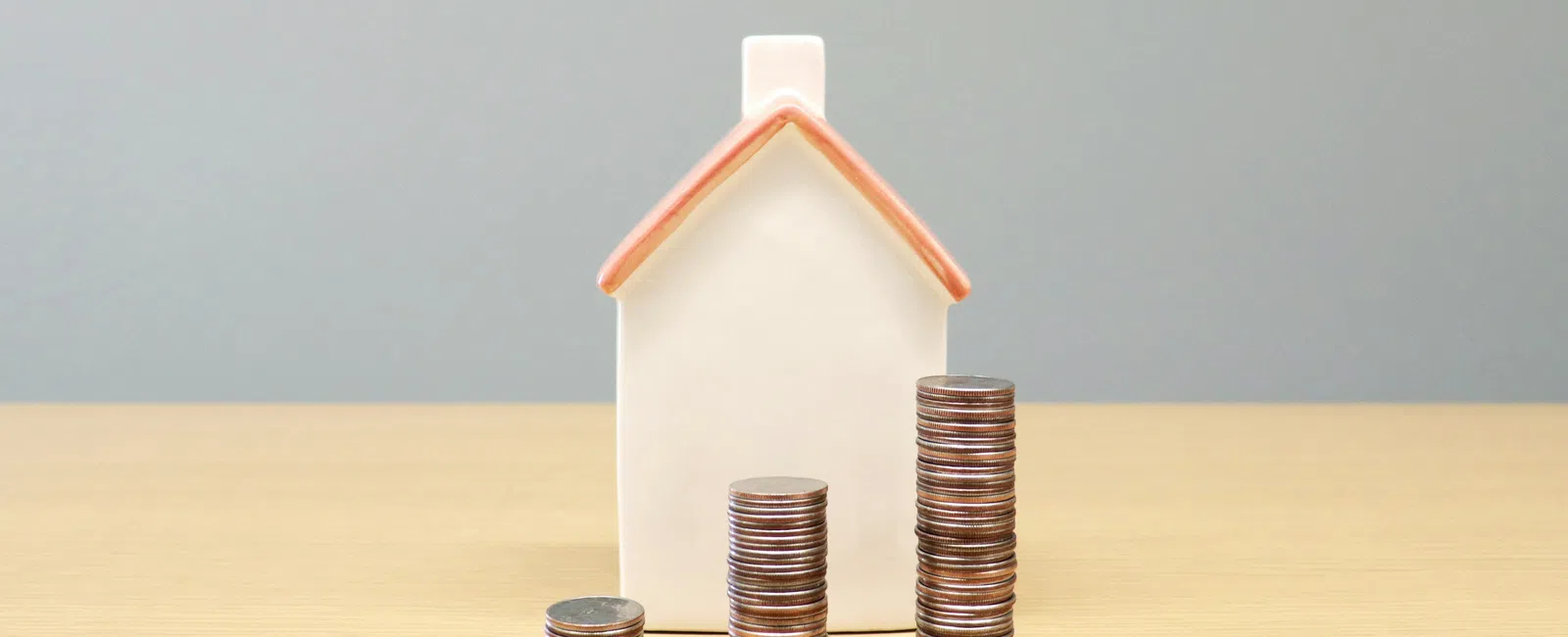 Three stacks of coins of varying heights in front of a small white house figurine with a red roof on a wooden surface.