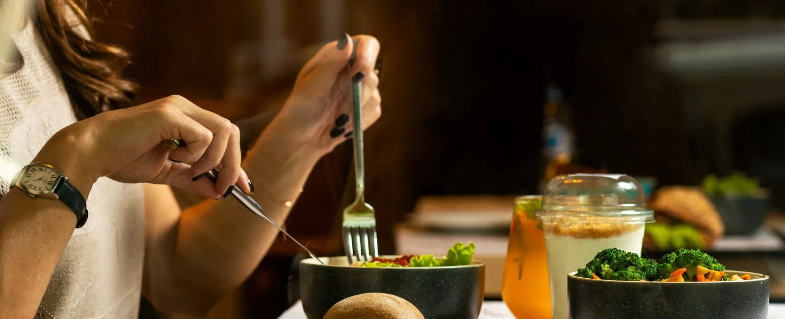Person eating salad with fork and knife at a table set with bread roll, drinks, and vegetable bowls in warm lighting