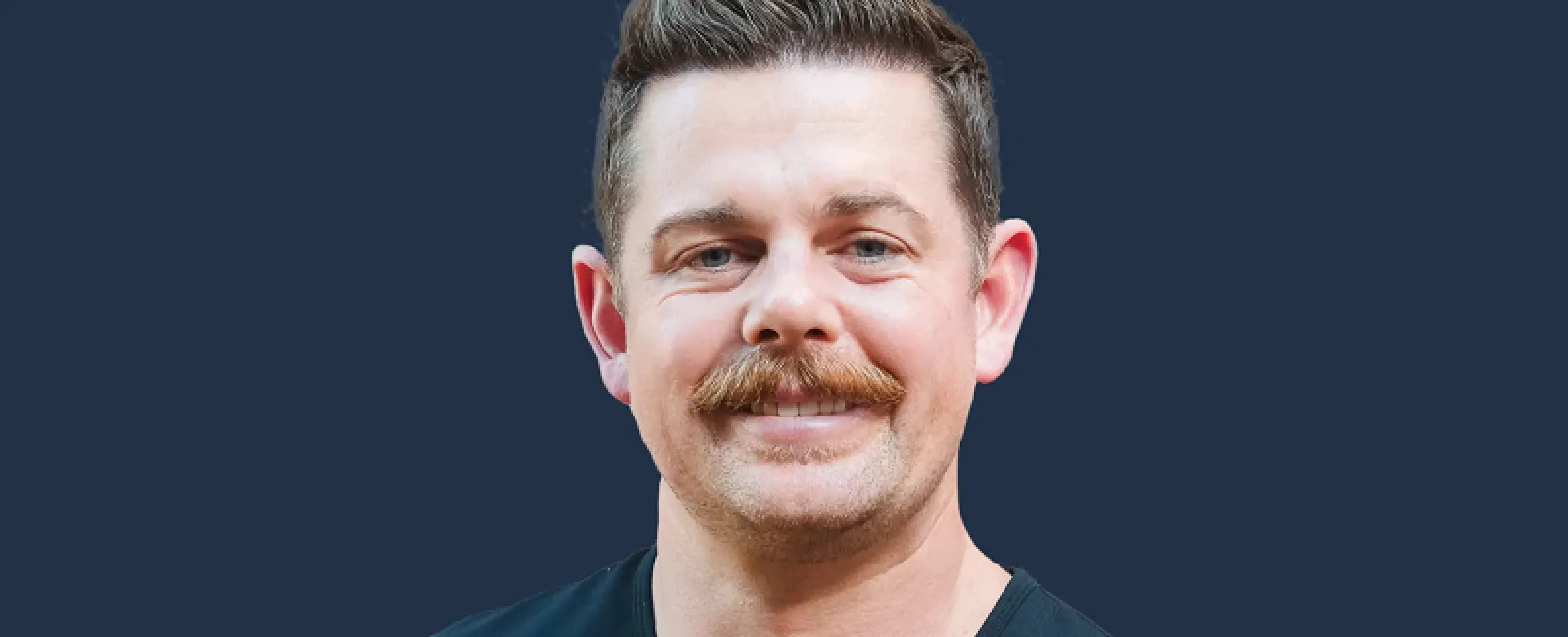 Portrait of a smiling man with a mustache and short hair wearing a black shirt against a dark blue background