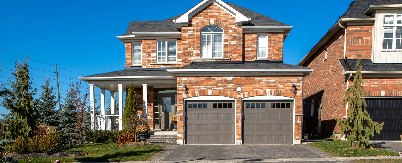 Two-story brick house with double garage doors and a blue recycling bin on the driveway under clear blue sky