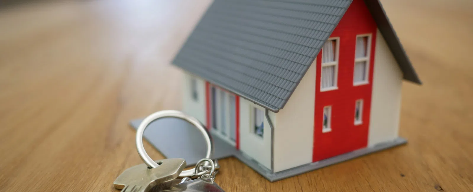 Key and house-shaped keychain placed next to a small model house on a wooden surface indoors.