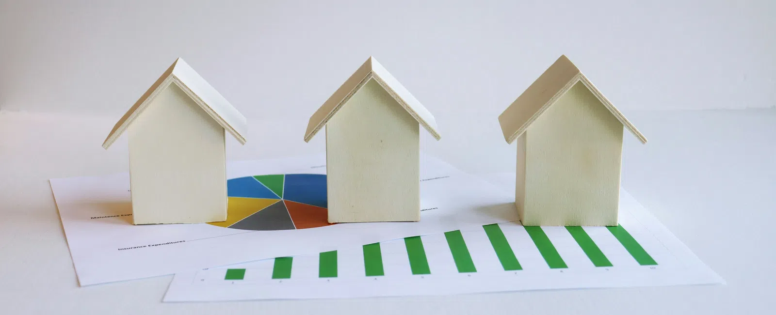 Three wooden house models on real estate financial charts including pie and bar graphs on a white surface
