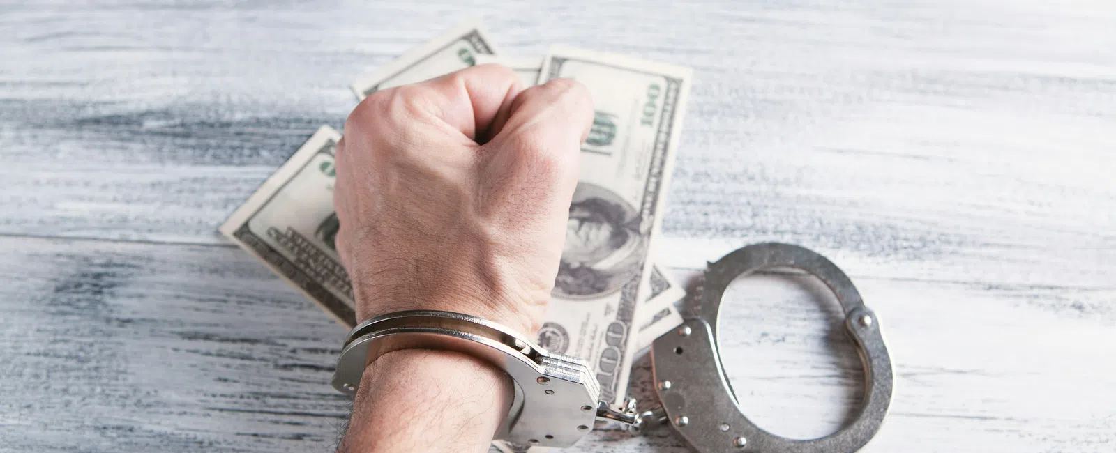 Handcuffed hand clenched in fist with US dollar bills on a wooden surface symbolizing financial crime.