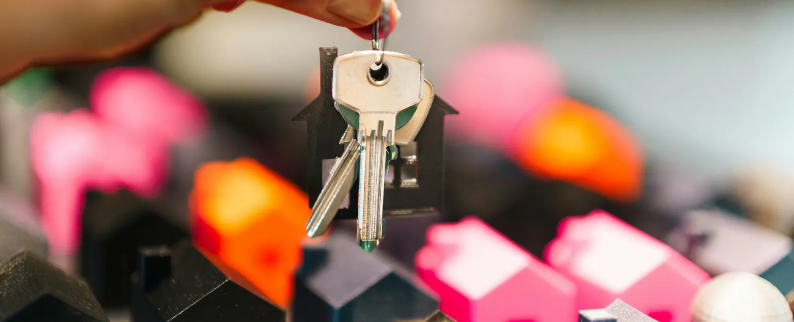 Hand holding house-shaped keyring with keys over colorful miniature house models representing real estate.