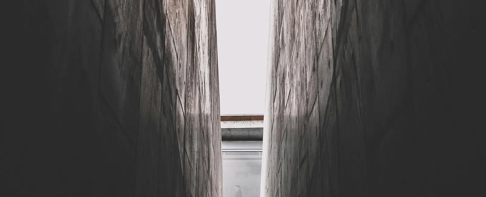 Narrow concrete alley with tall walls and dark shadows leading to a staircase with handrails and an opening above.