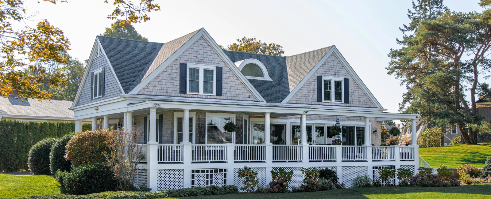 Large white house with wraparound porch, grey shingle roof, and lush green lawn on a sunny day