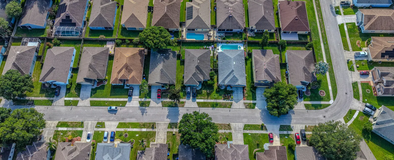 Aerial view of a suburban neighborhood with rows of houses, green lawns, trees, and parked cars on streets.