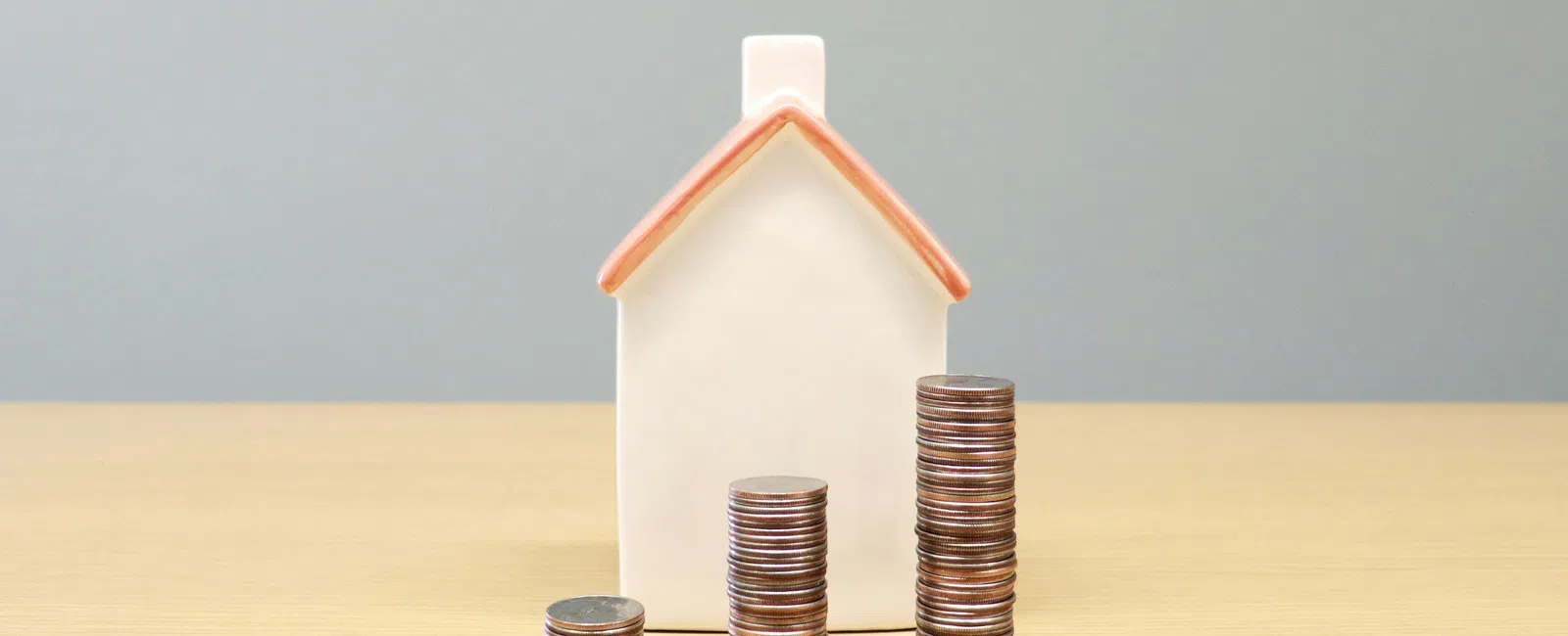 Stacked coins of increasing height in front of a white house model on wooden table with grey background.