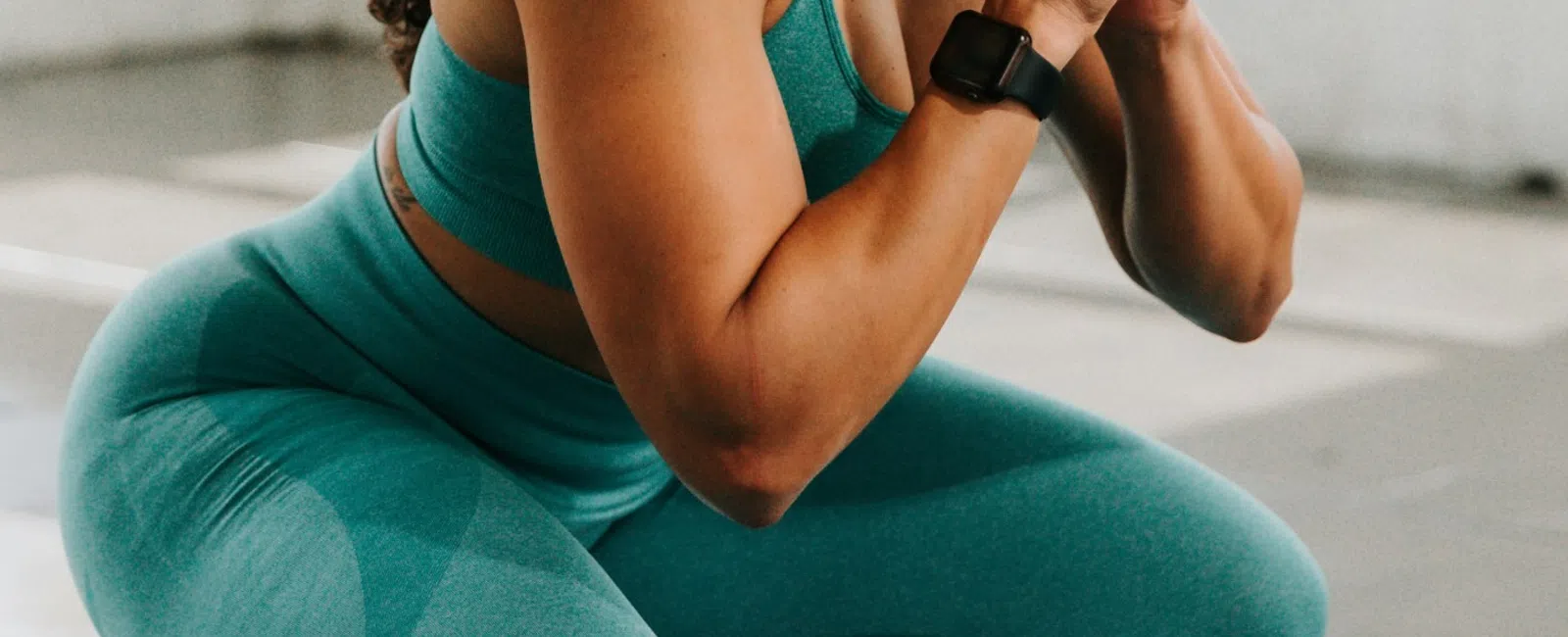 Fit woman with curly hair in teal workout clothes performing a squat exercise indoors by large windows.