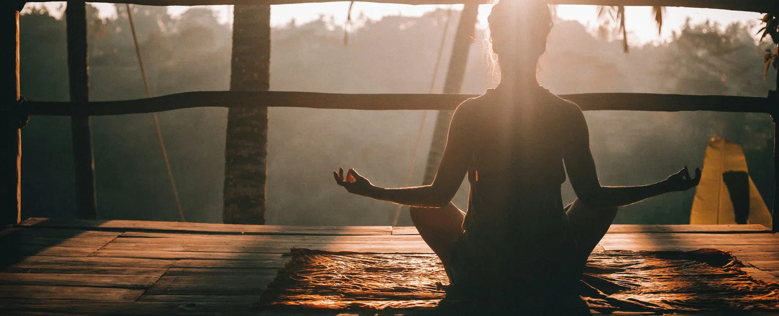 Silhouette of person meditating in lotus pose on wooden deck at sunrise with palm trees in background.