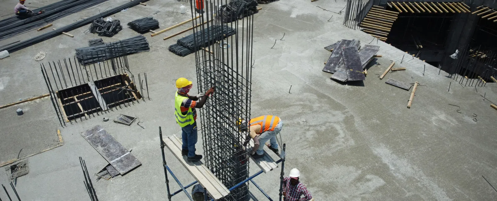 Construction workers assembling steel reinforcement bars on a large concrete building site under clear daylight.