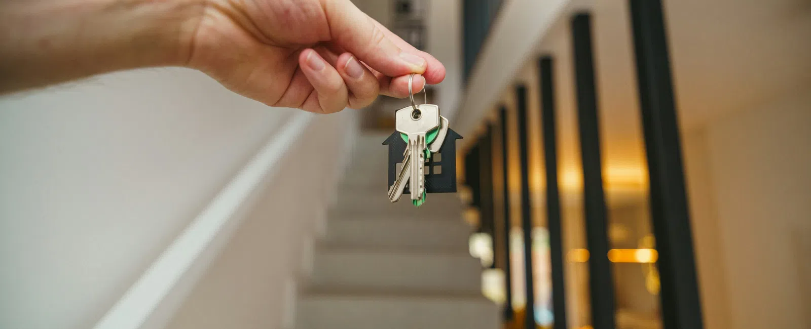 Hand holding keys with a house-shaped keychain in front of a modern home's staircase and interior.