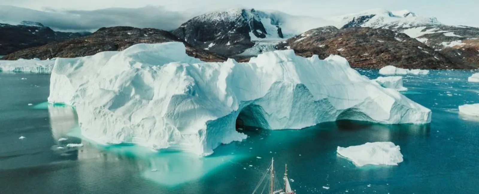 Sailboat near a large ice arch floating iceberg with mountainous snowy landscape in the background