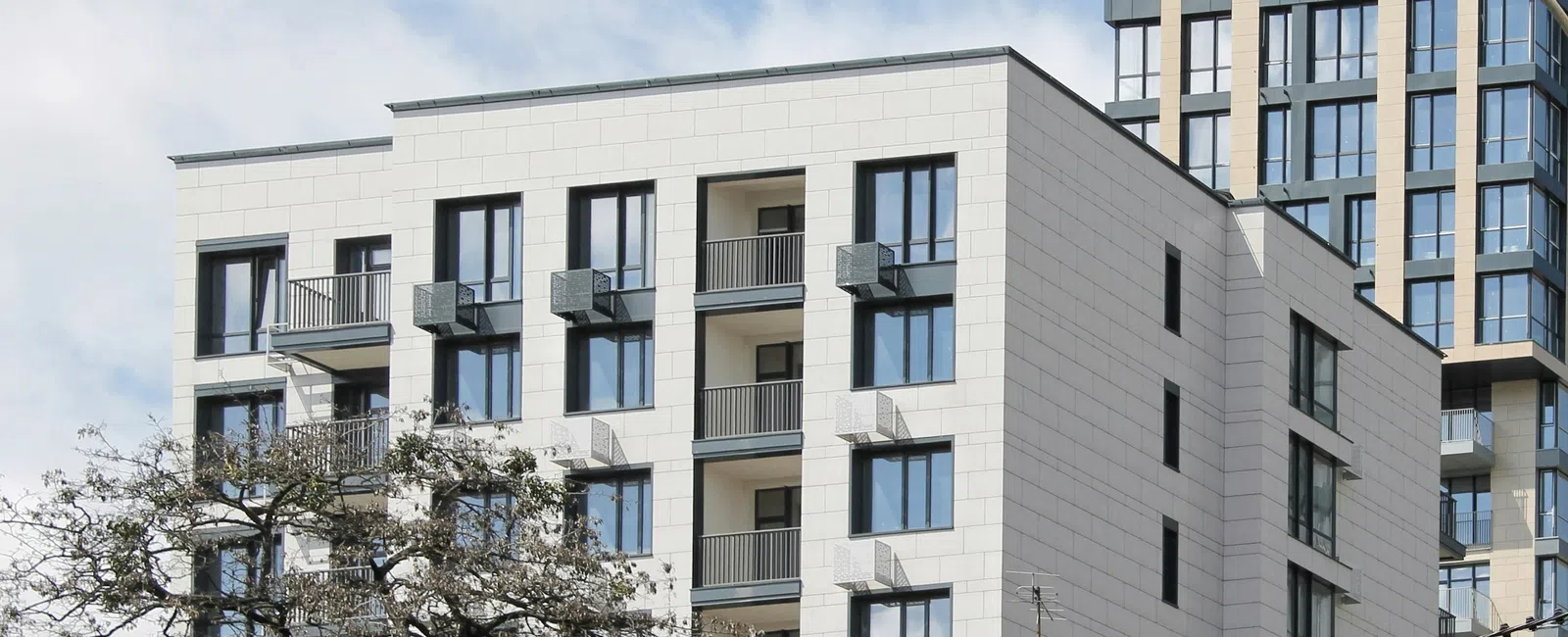 Modern white residential building with balconies and large windows under partly cloudy sky.