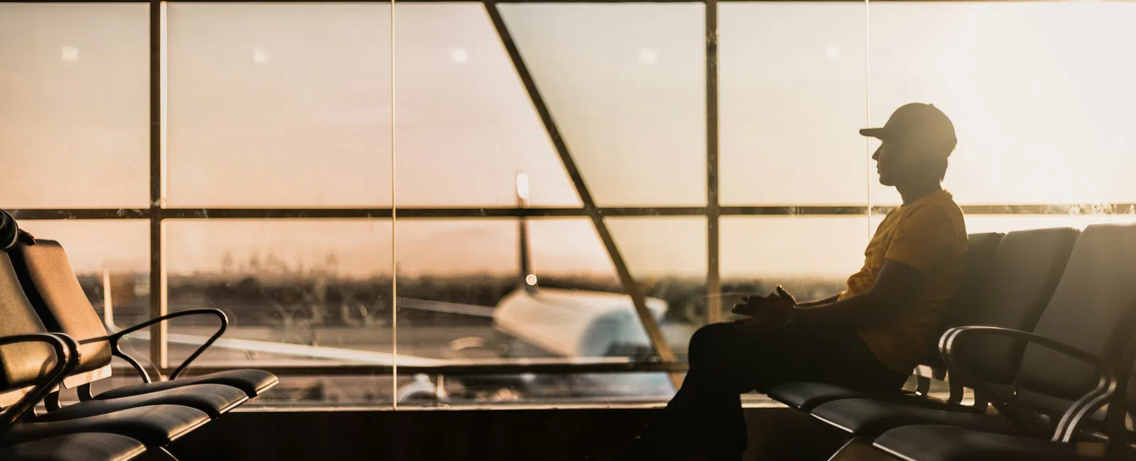 Silhouette of a man sitting in an airport lounge with a plane visible through large glass windows at sunset.