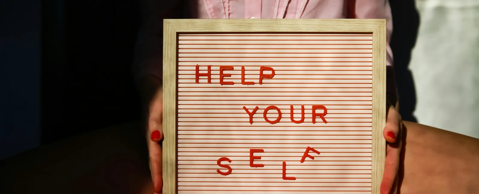 Person holding letter board with red text 'HELP YOURSELF' in sunlight with shadows and pink shirt.