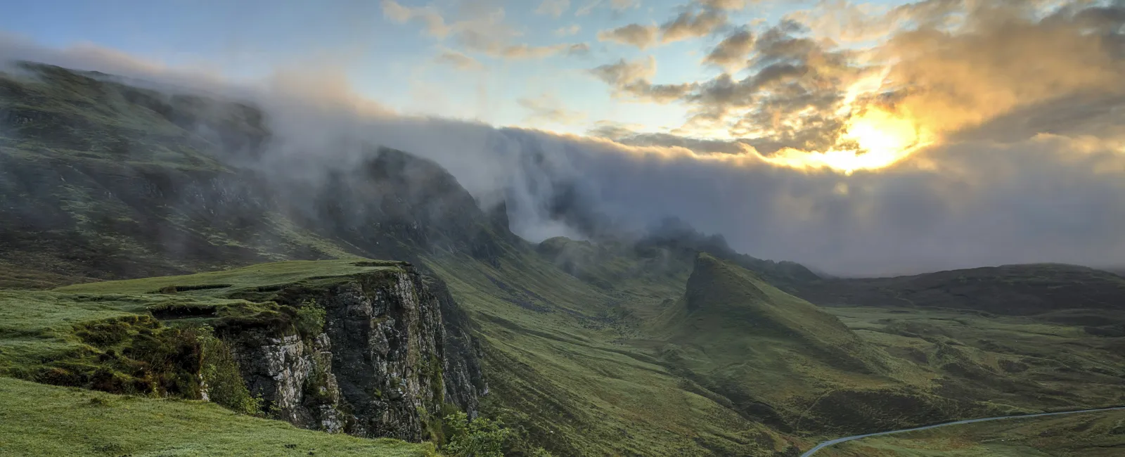 Sunset over misty green cliffs and winding road in a mountainous landscape under a cloudy sky