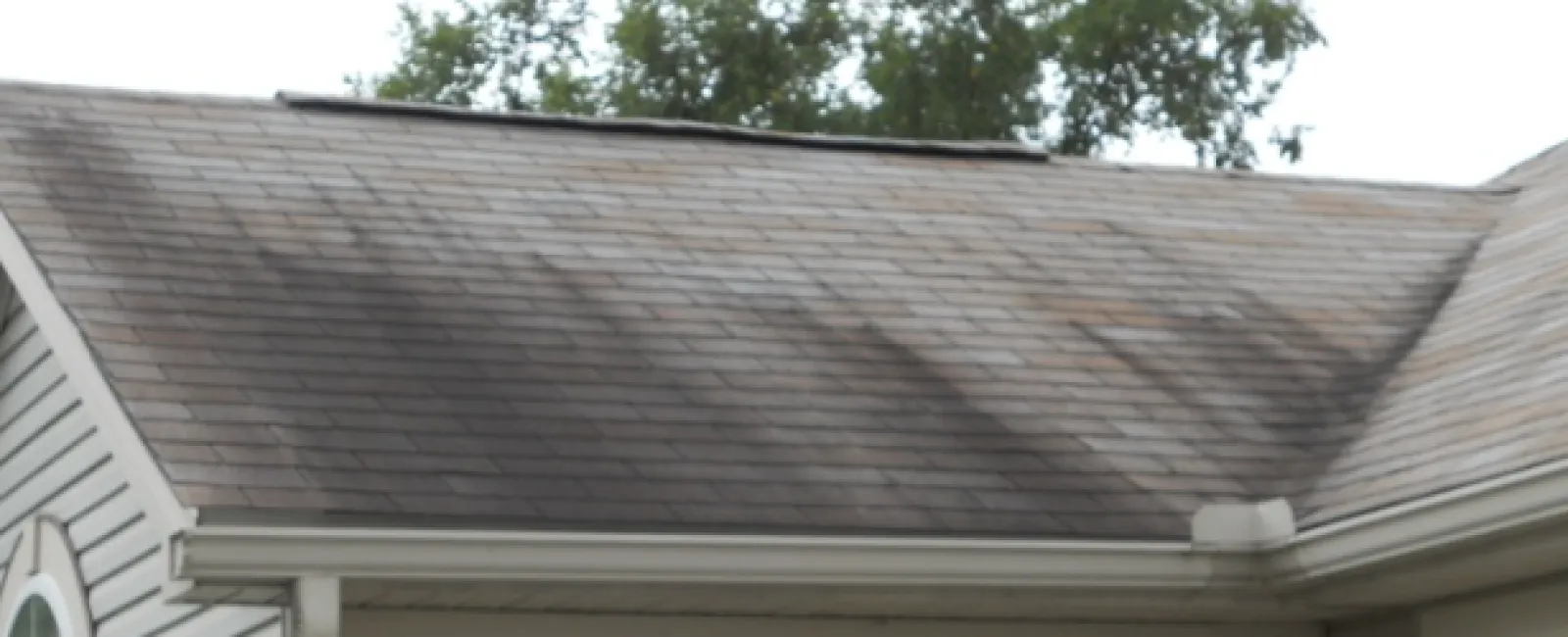 Close-up of a house roof with asphalt shingles and white siding under a cloudy sky.