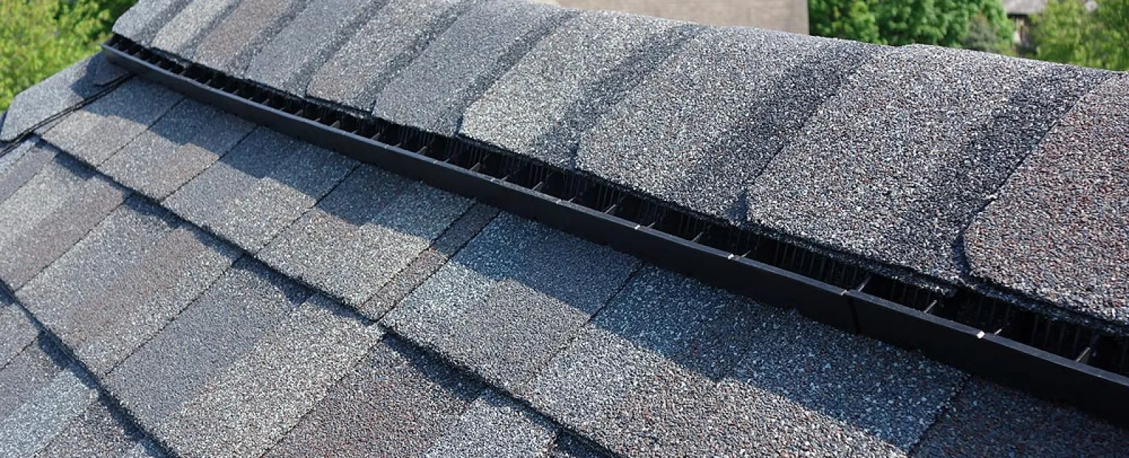 Close-up of a gray composite shingle roof with ridge vent installed under a clear sky in a residential area.