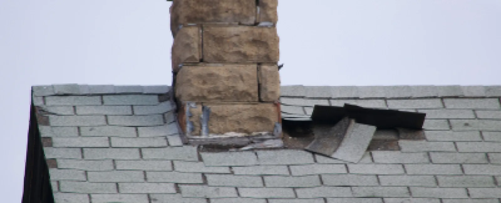 Roof with gray shingles and stone chimney showing damaged and missing shingles near the chimney base.