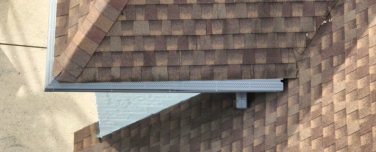 Aerial view of a brown shingle roof with gutter system and a downspout over a concrete ground.