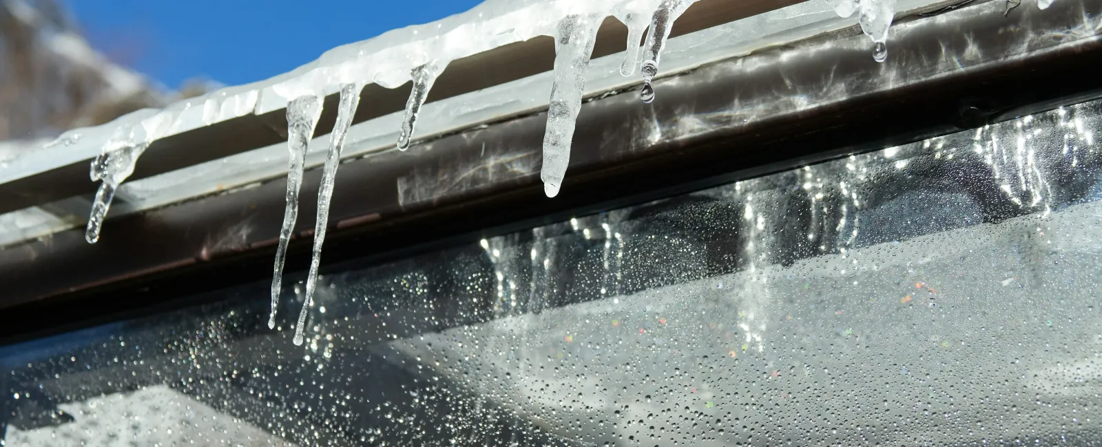 Icicles hanging from a roof edge over a glass surface with water droplets under a clear blue sky.