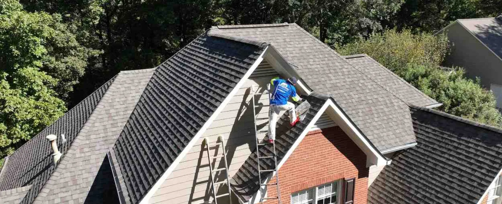 Worker on ladder painting exterior of brick and siding house surrounded by trees on sunny day