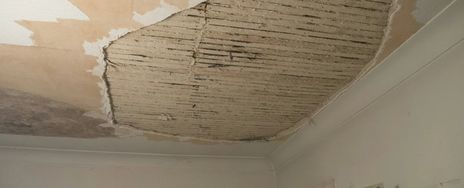 Peeling damaged ceiling plaster exposing wooden laths in a room with shelves and radio below.
