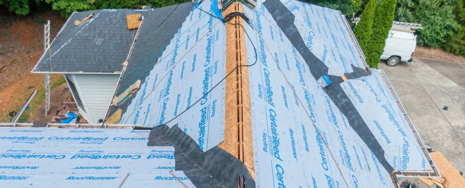 A residential roof under construction with waterproof underlayment partially covered by black shingles surrounded by trees.