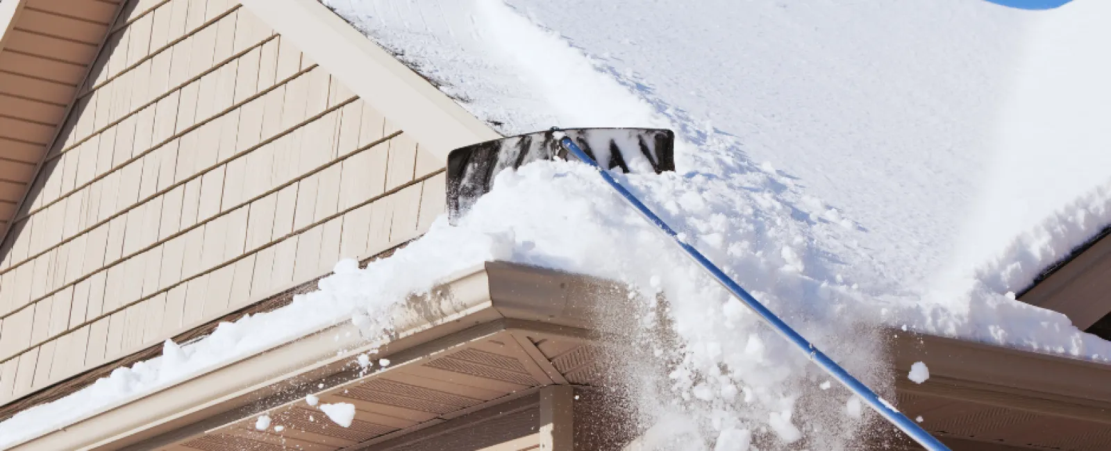 Person using a long snow shovel to clear heavy snow off a beige house roof on a clear winter day.