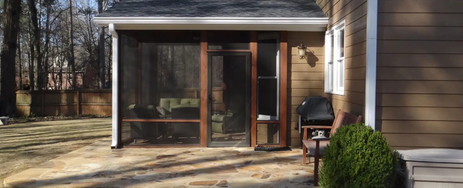 Stone patio with screened porch, outdoor grill, bench, and brown siding house surrounded by trees and yard.