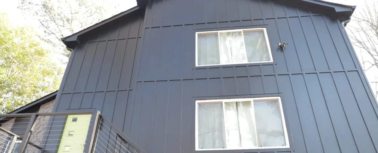 Modern dark gray house exterior with two large windows and a balcony with metal railing under bright daylight.