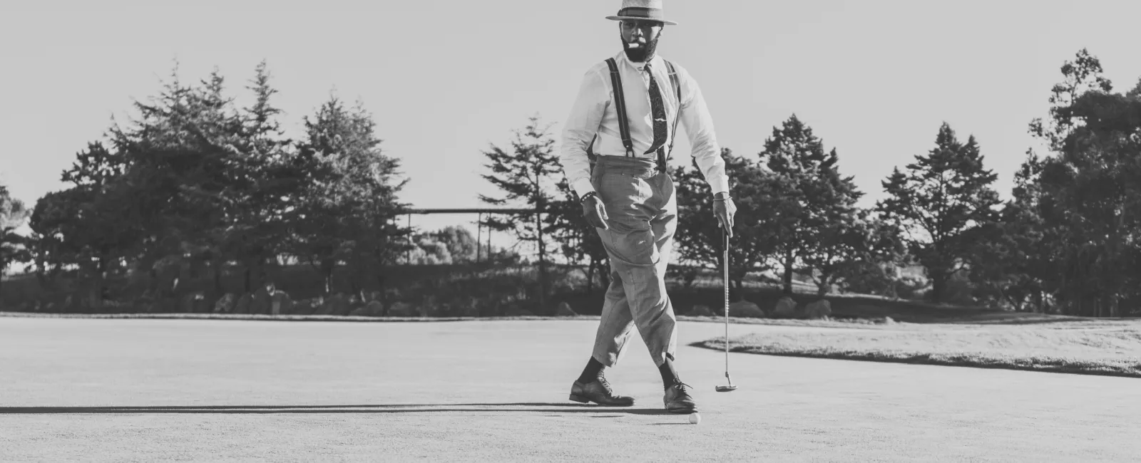 Black and white image of a man in vintage attire playing golf on a sunny course with trees in the background.