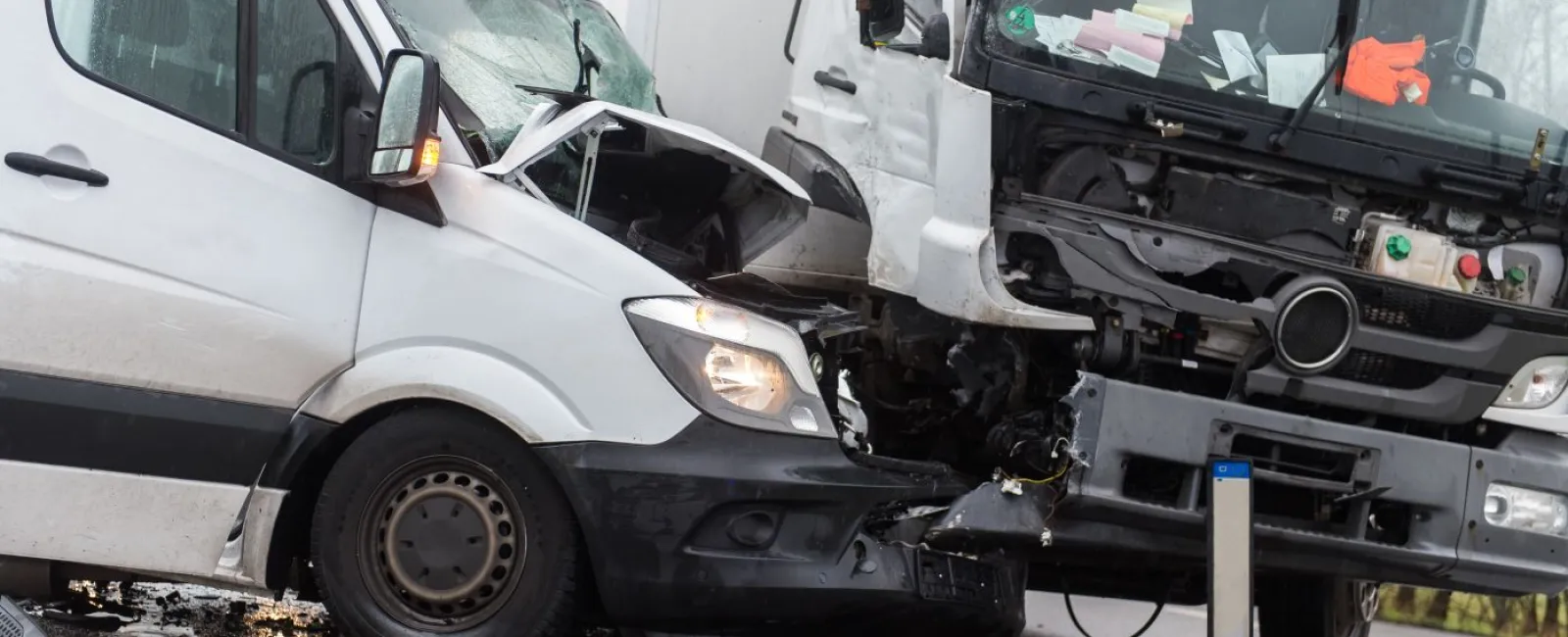 White van and truck with severe front-end damage after collision on wet road with debris scattered.