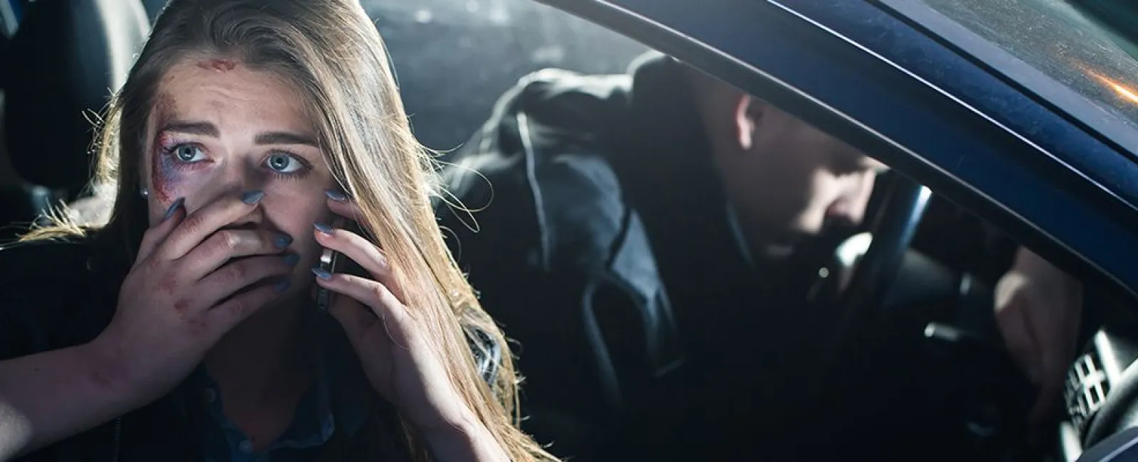 Distressed woman with a bruised face makes a phone call from a car while a man sits in the driver's seat.