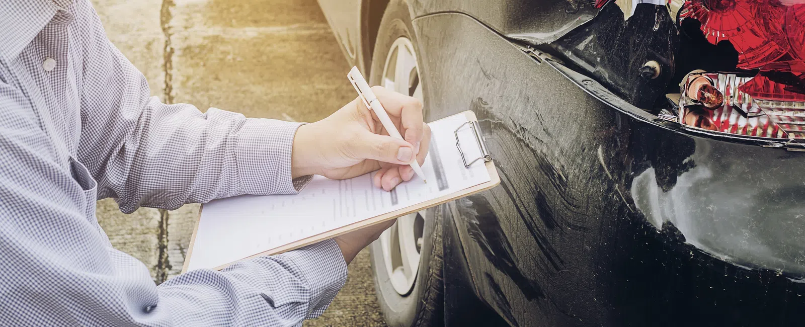 Person inspecting and documenting damage on rear side of black car with clipboard and pen outdoors.